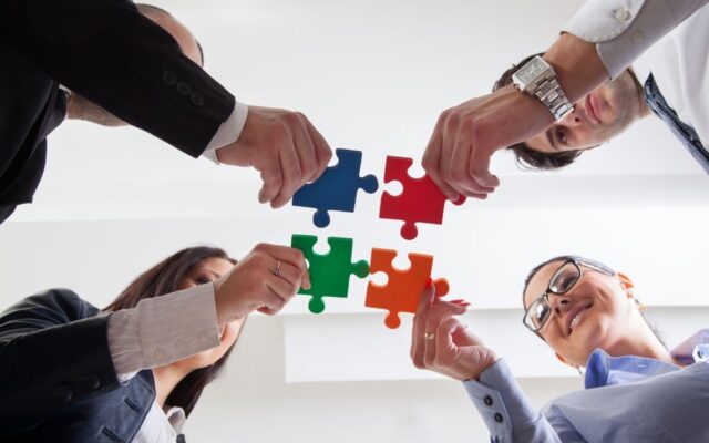 Four professionals collaborate to connect puzzle pieces against a white background, symbolizing teamwork, problem-solving, and the joining of diverse ideas in a corporate environment while maintaining their mental health.