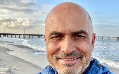 A smiling man with a shaved head, rejuvenating his energy level, enjoys a bright day at the beach, wearing a blue jacket, with a wooden pier in the background and waves gently breaking on the