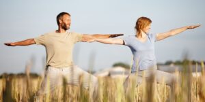 A man and woman improving their well-being by practicing yoga together in a sunny, open field, performing a standing pose with arms stretched out to the sides, embodying balance and tranquility amidst nature.